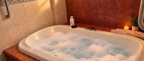 A bathtub filled with bubbles, a handrail, a bottle of soap, and a candle on a bathroom counter.