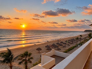 A beach with a sunset, umbrellas, and palm trees. - Costa Bonita Beachfront Family Condo (Mazatlán)