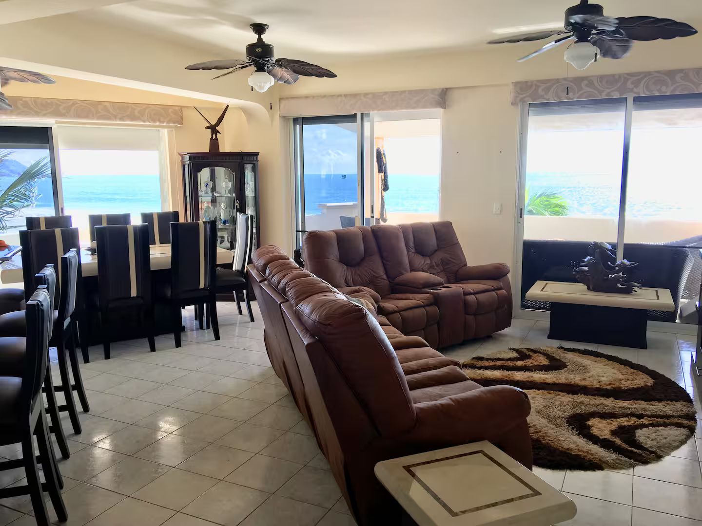 A living room with a brown leather sofa, a dining table with chairs, a ceiling fan, and a view of the ocean through large windows.