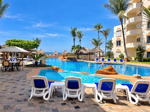A resort pool area with lounge chairs, palm trees, and a clear blue sky. - Costa Bonita Beachfront Family Condo (Mazatlán)