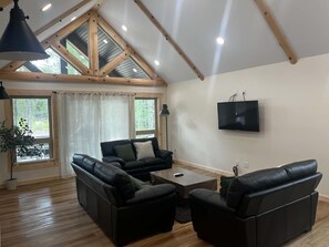 Ein modernes Wohnzimmer mit geneigter Decke, Holzbalken, einem Flachbildfernseher, Ledersofas und einer Topfpflanze.
