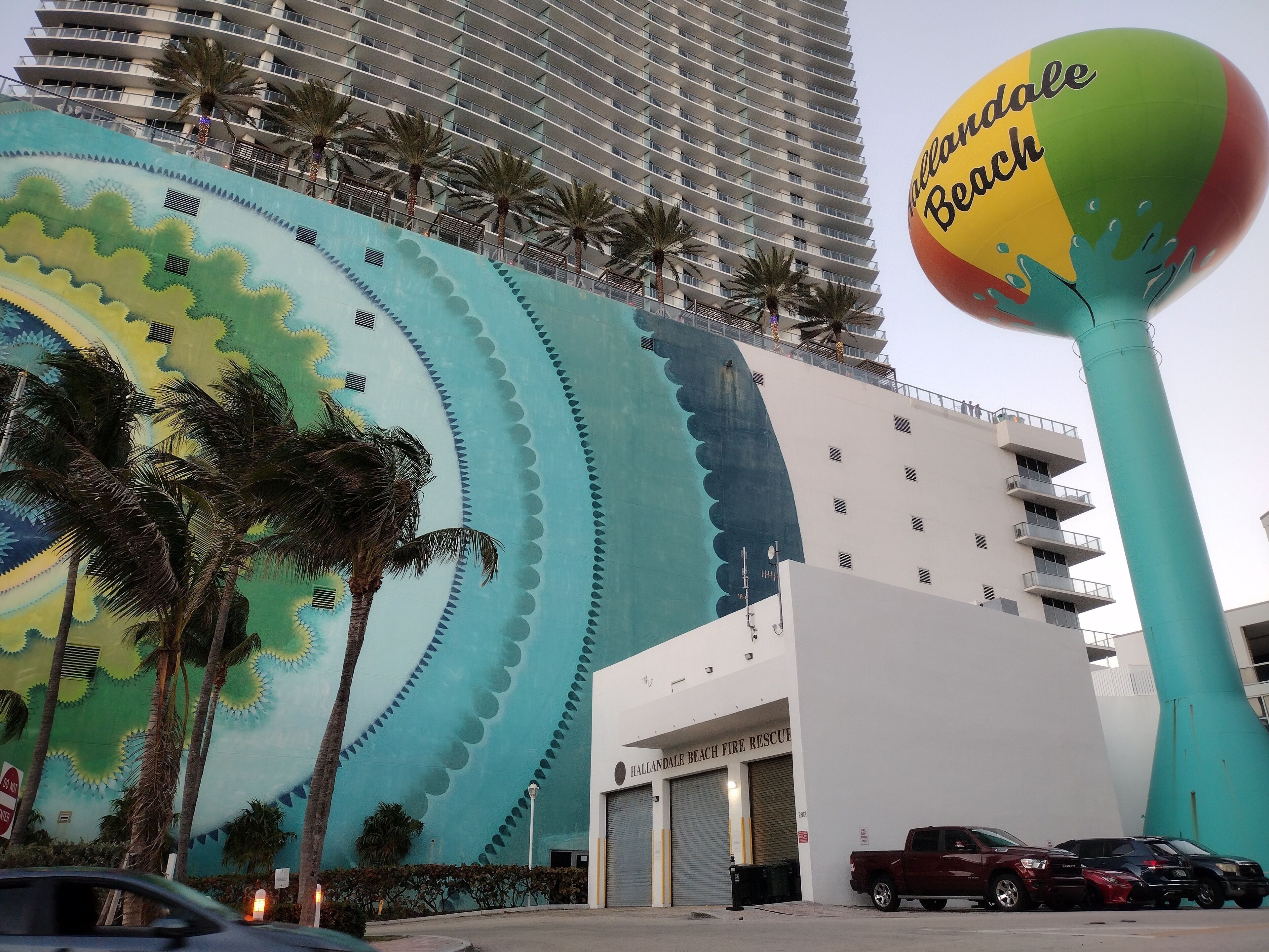 A building with a large mural of palm trees and a beach scene, a fire rescue station, and parked vehicles.