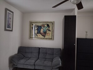 Une pièce avec un canapé gris, un ventilateur de plafond, un tableau abstrait encadré et une porte fermée.