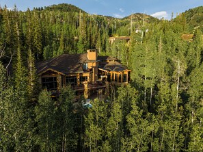 A large, multi-story lodge surrounded by dense forest, with a prominent chimney and multiple windows. - Ski-In/Ski-Out 6BR Luxury Estate with Indoor Pool, Hot Tub & Theater at The Colony, Sleeps 26 (Park City)