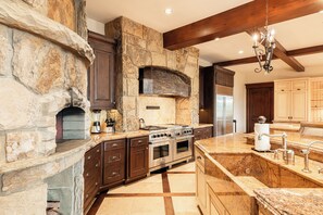 A kitchen with a stone fireplace, wooden cabinets, a built-in oven, and a large island with a sink. - Ski-In/Ski-Out 6BR Luxury Estate with Indoor Pool, Hot Tub & Theater at The Colony, Sleeps 26 (Park City)