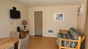 A living room with a wooden dining table, chairs, a sofa, a television mounted on the wall, a lamp, and a door leading to another room. - 2 Queen Beds, 1 Sofa Bed, Kitchen. No Pets Allowed (Packwood)