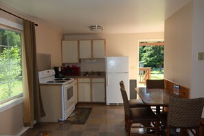 Een kleine keuken met een fornuis, magnetron en koelkast, een eettafel met stoelen, en uitzicht op groen buiten.