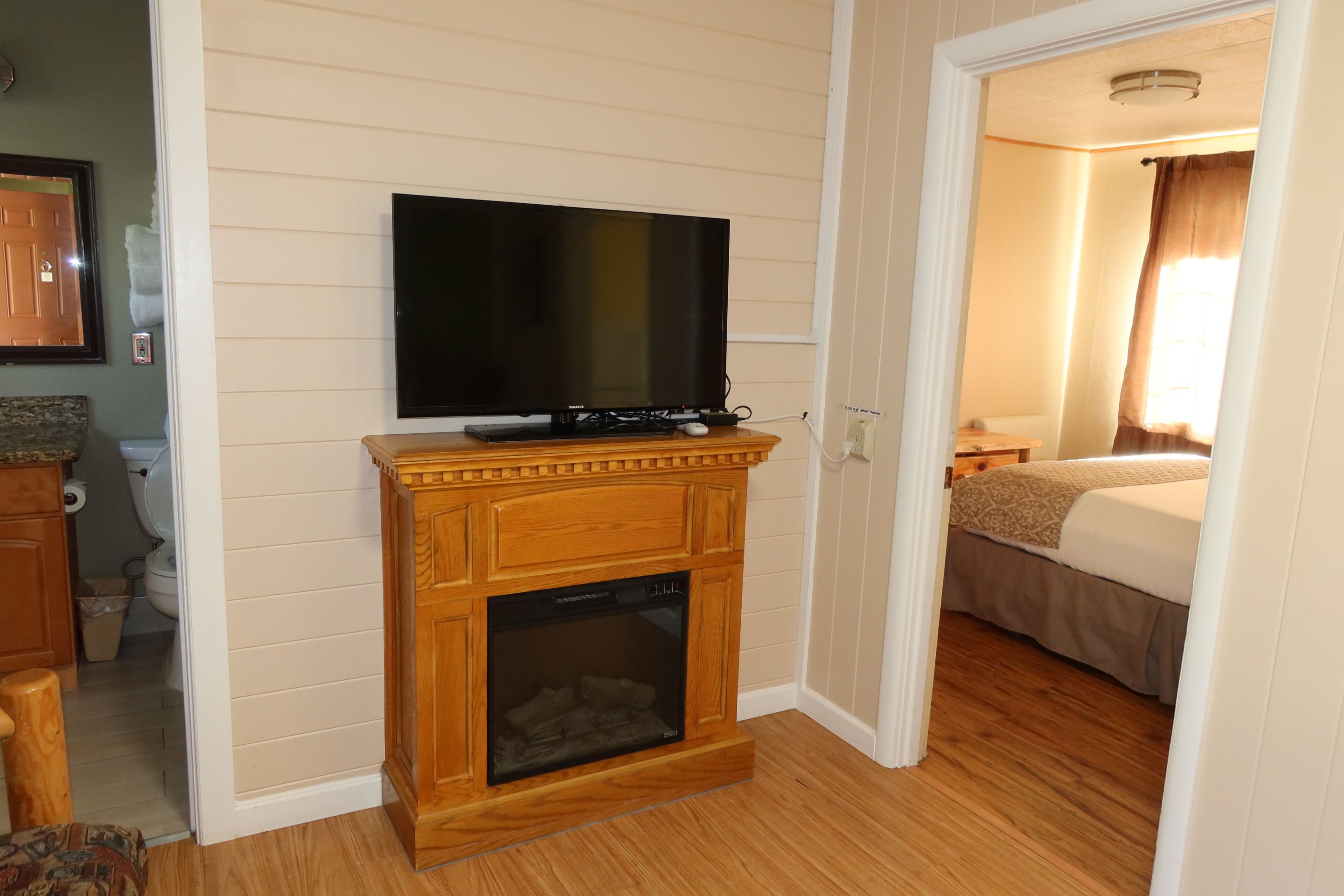 A hotel room with a wooden fireplace, a television mounted above it, a bed, and a bathroom visible in the corner.