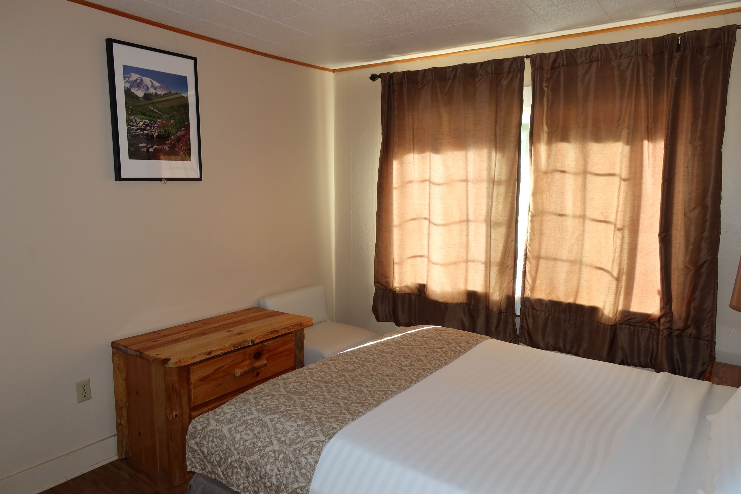Un dormitorio con una cama, una mesita de noche de madera, un cuadro enmarcado en la pared y una ventana con cortinas.