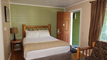 A single bed with a wooden headboard, a bedside table with a lamp, a patterned chair, and a door with a number plate.
