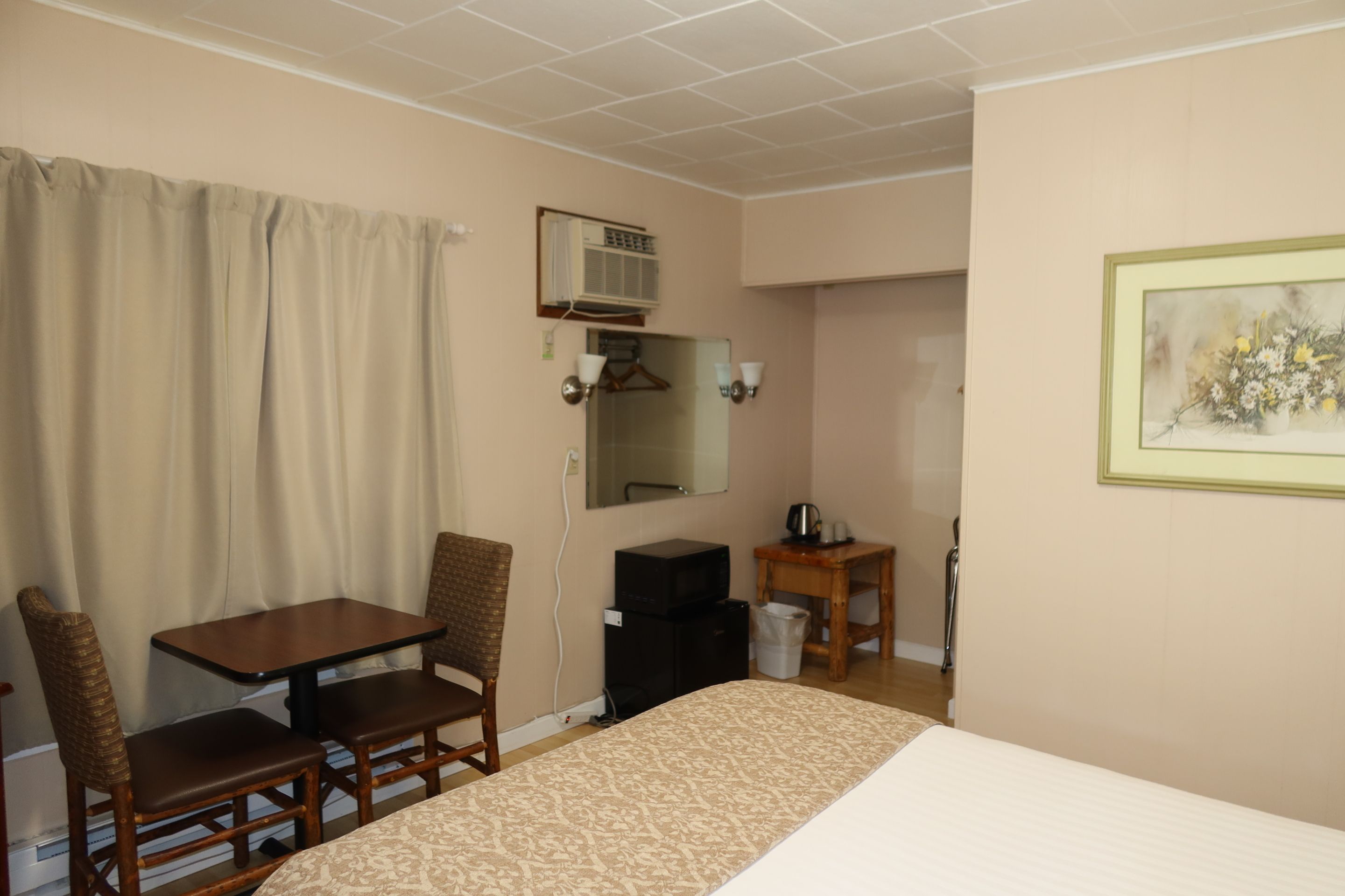 Une chambre d’hôtel équipée d’un lit, d’un bureau, d’une chaise, d’une petite table, d’un miroir, d’un tableau au mur et d’un appareil de climatisation.