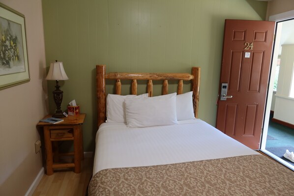 A room with a wooden headboard bed, a bedside table with a lamp, and a door with a number.