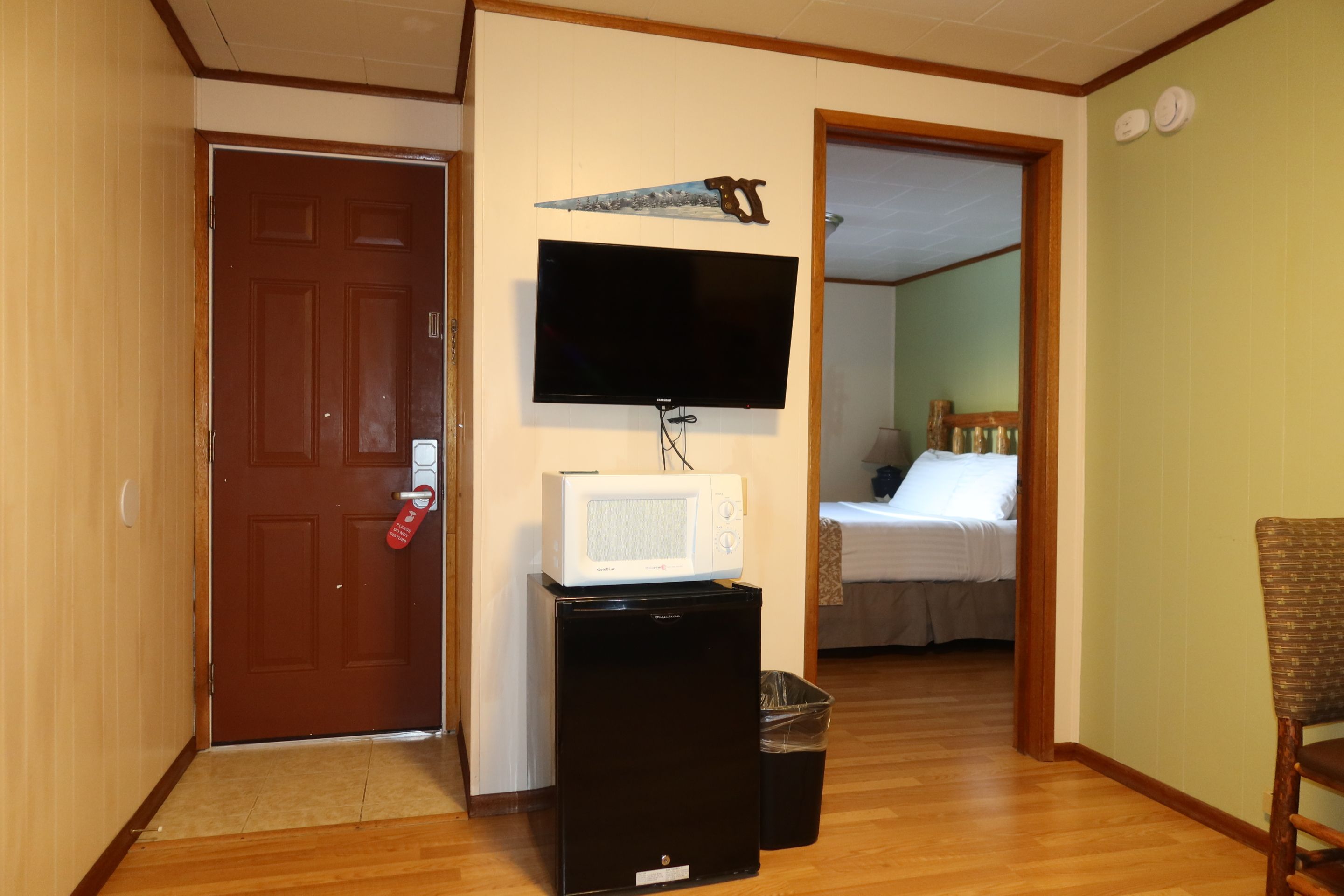 Een kamer met een aan de muur bevestigde televisie, een magnetron op een klein tafeltje, een deur met een rood bordje en een bed op de achtergrond.