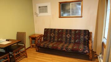 A room with a wooden floor, a sofa with a patterned cover, a small wooden table, a chair, and a window with blinds.