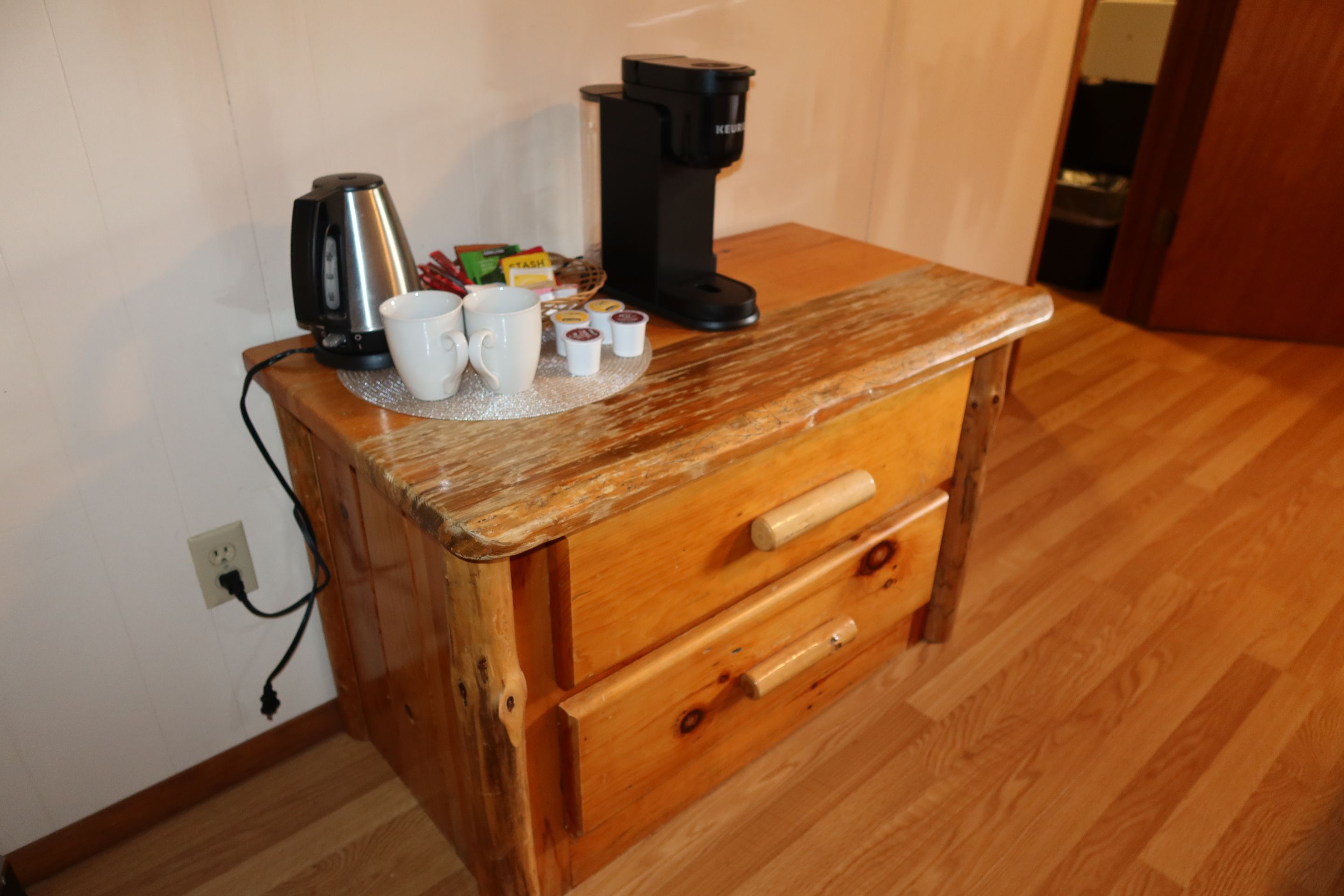 Un meuble en bois avec un plan de travail, une bouilloire, une machine à café et plusieurs dosettes de café.