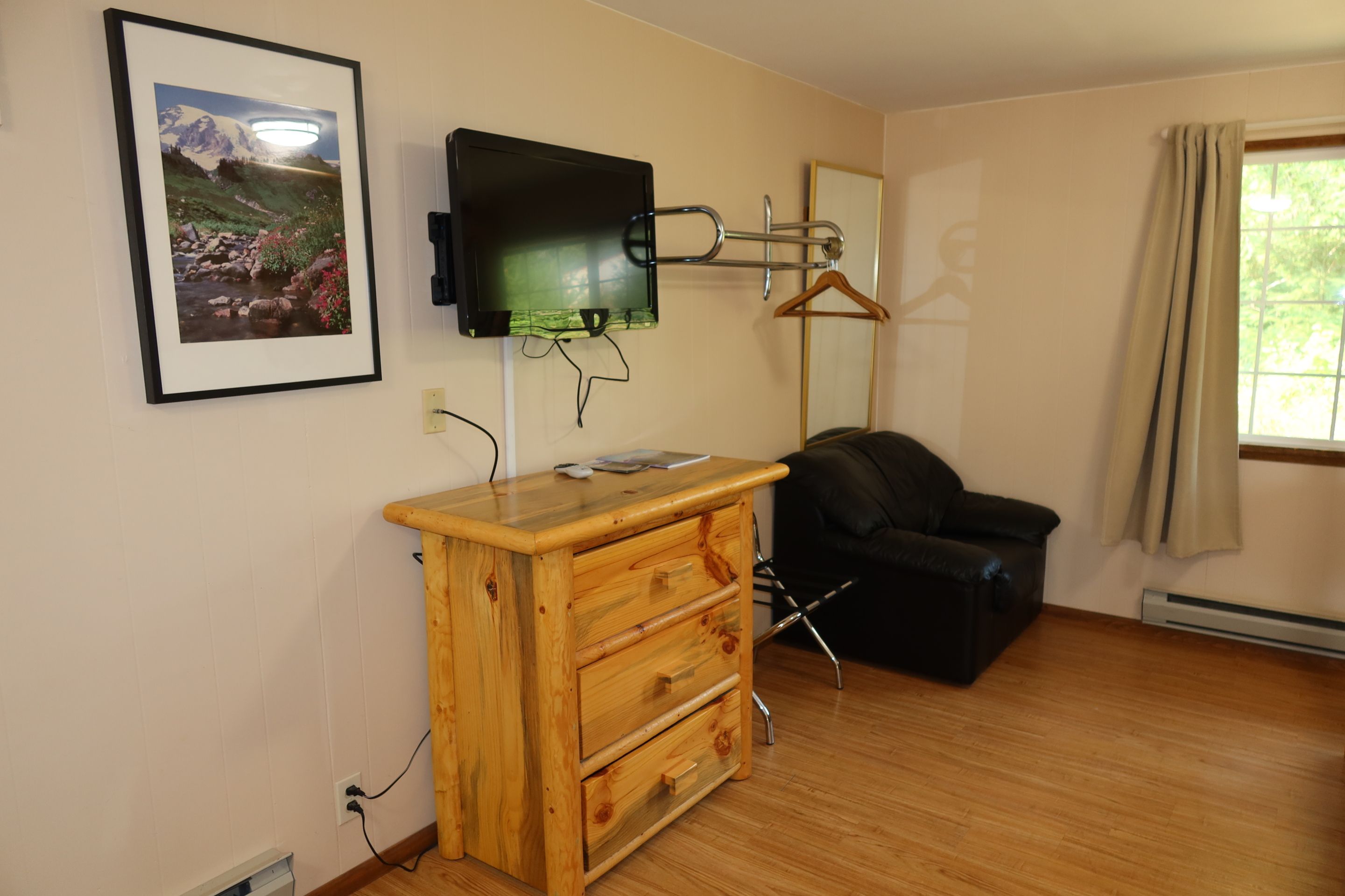 Une pièce comprenant une commode en bois, un fauteuil en cuir noir, un téléviseur fixé au mur et un tableau représentant un paysage.