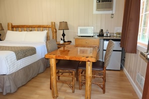 Una habitación con cama, mesa de madera, sillas, microondas, refrigerador y una ventana con cortinas.