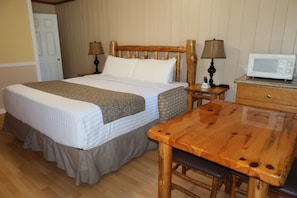 Una habitación de hotel con cama, cabecero de madera, dos mesitas de noche con lámparas, microondas, mesa de madera y un banco.