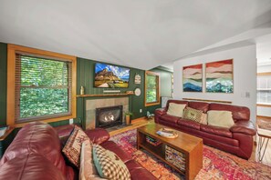 A living room with a fireplace, a large flat-screen TV, a red leather sofa, and a colorful rug. - The Barking Beech: Pet Friendly, Hot Tub, Firepit, Fenced Yard, 4 TVs! (Banner Elk)
