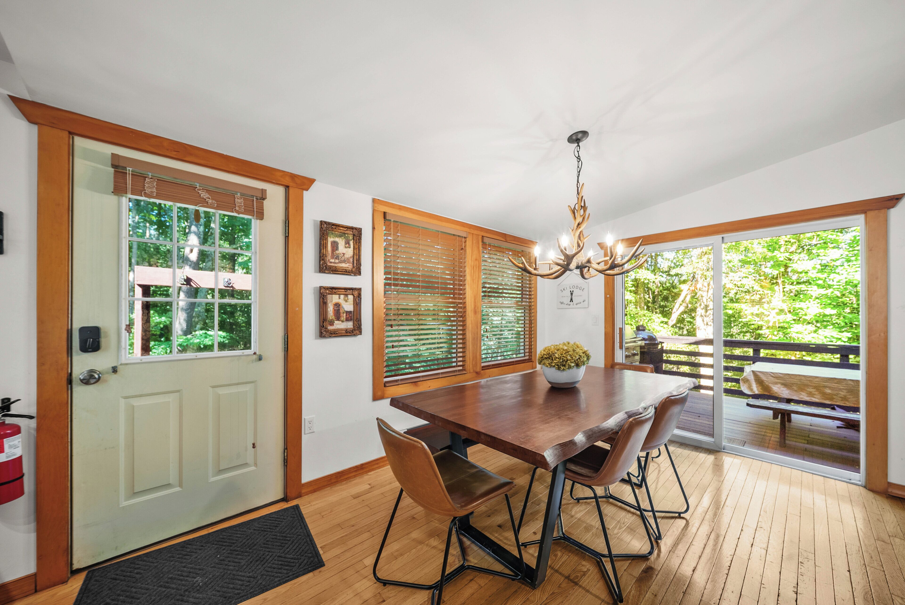 A dining area with a wooden table, chairs, and a chandelier. There are windows with blinds, a door, and a view of the outdoors.