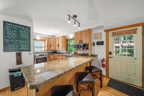 A kitchen with granite countertops, wooden cabinets, a microwave, and a bar area with stools. - The Barking Beech: Pet Friendly, Hot Tub, Firepit, Fenced Yard, 4 TVs! (Banner Elk)