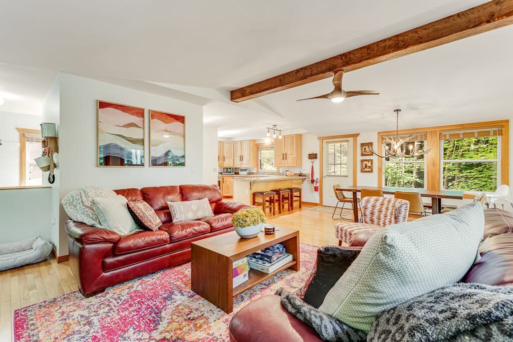 A spacious living room with a red leather sofa, a coffee table, and a patterned rug. There is a dining area with wooden chairs and a table, and a kitchen in the background.