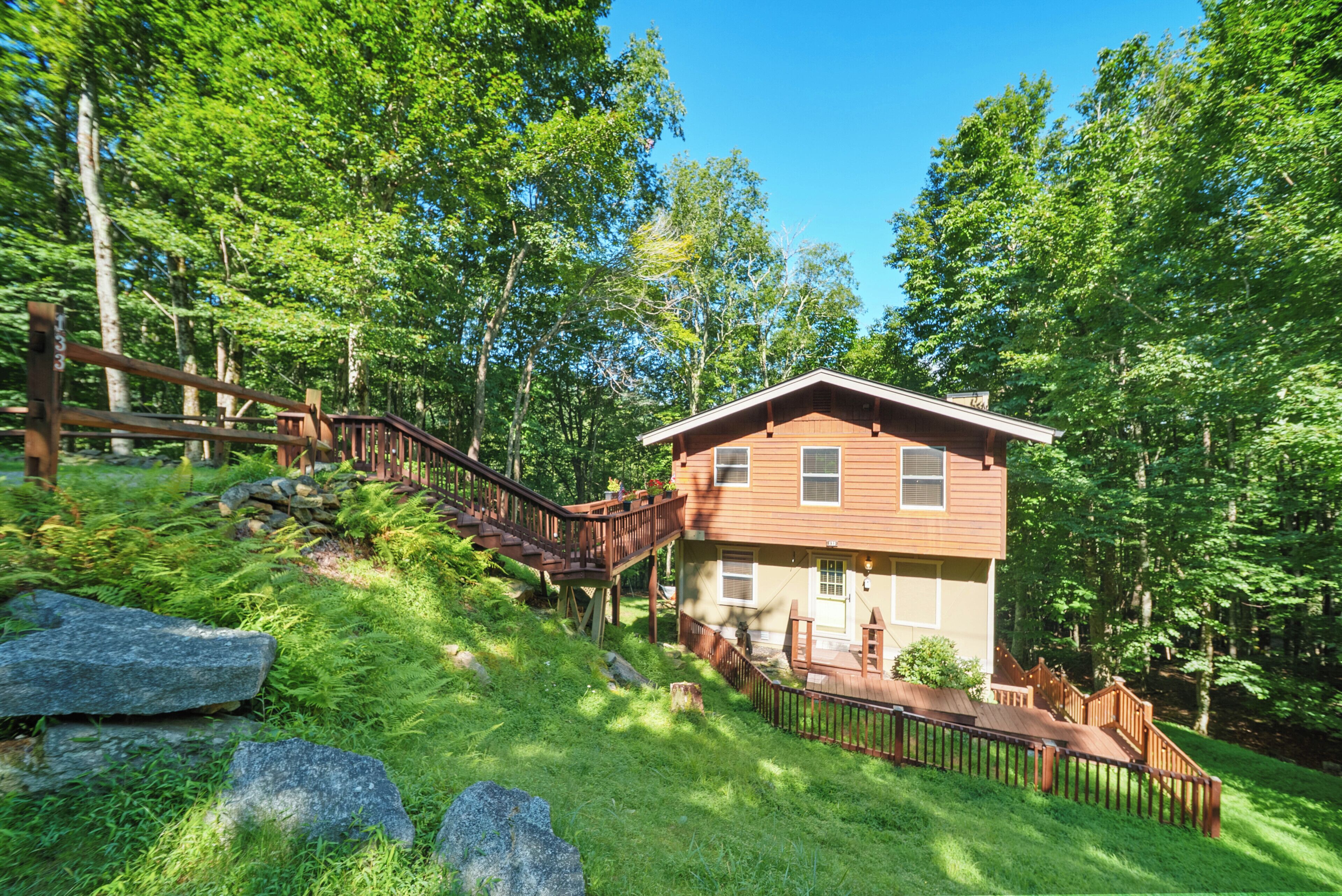 A wooden cabin with a deck and stairs, surrounded by lush greenery and trees.
