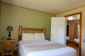 Un dormitorio con cabecera de madera, una cama con ropa de cama blanca, una mesita de noche con lámpara y una puerta que da a otra habitación.