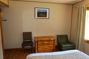 A room with a bed, a wooden nightstand, a green chair, a wooden chair, and a framed picture on the wall.