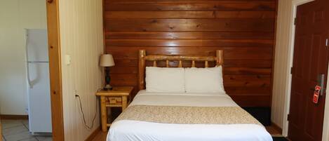 Een kamer met een bed met houten hoofdbord, een nachtkastje met een lamp, een deur met een rode hendel, en een koelkast in de aangrenzende kamer.