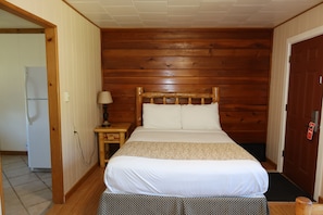 Una habitación con una cama de cabecera de madera, una mesita de noche con lámpara, una puerta con picaporte rojo y un refrigerador en la habitación contigua.