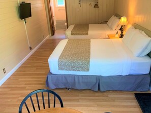 A hotel room with two beds, a TV mounted on the wall, a bedside table with a lamp, and a wooden floor.