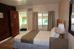 Una habitación de hotel con una cama, una silla, una lámpara, una puerta y un ventanal con cortinas.