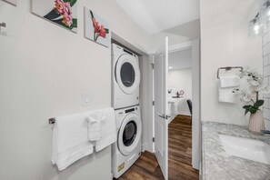 A modern bathroom with a double washer-dryer unit, a wooden floor, a marble sink, and a white orchid plant. - Chic, Upgraded 2BR with Stunning Ocean Views (Lahaina)