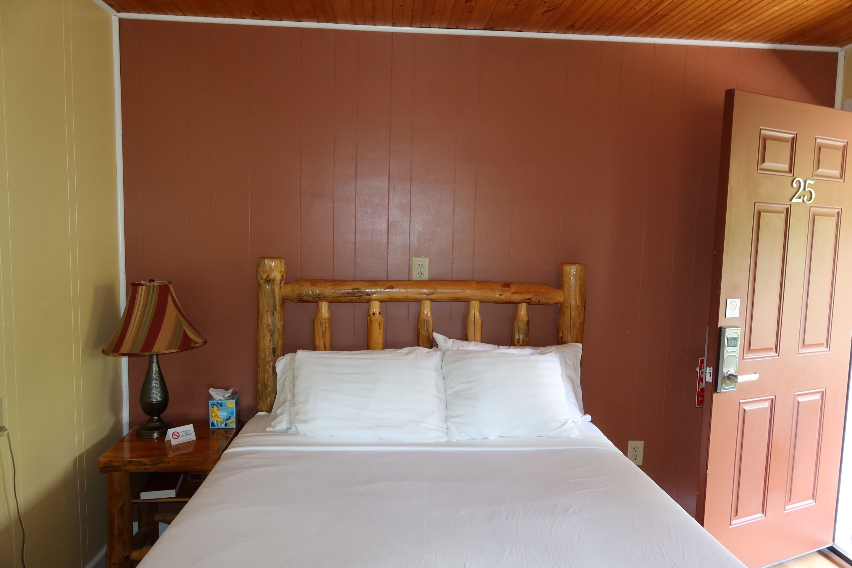 Un lit simple avec une tête de lit en bois, dans une pièce dont le mur est de couleur marron et dont la porte est marquée du numéro 25.