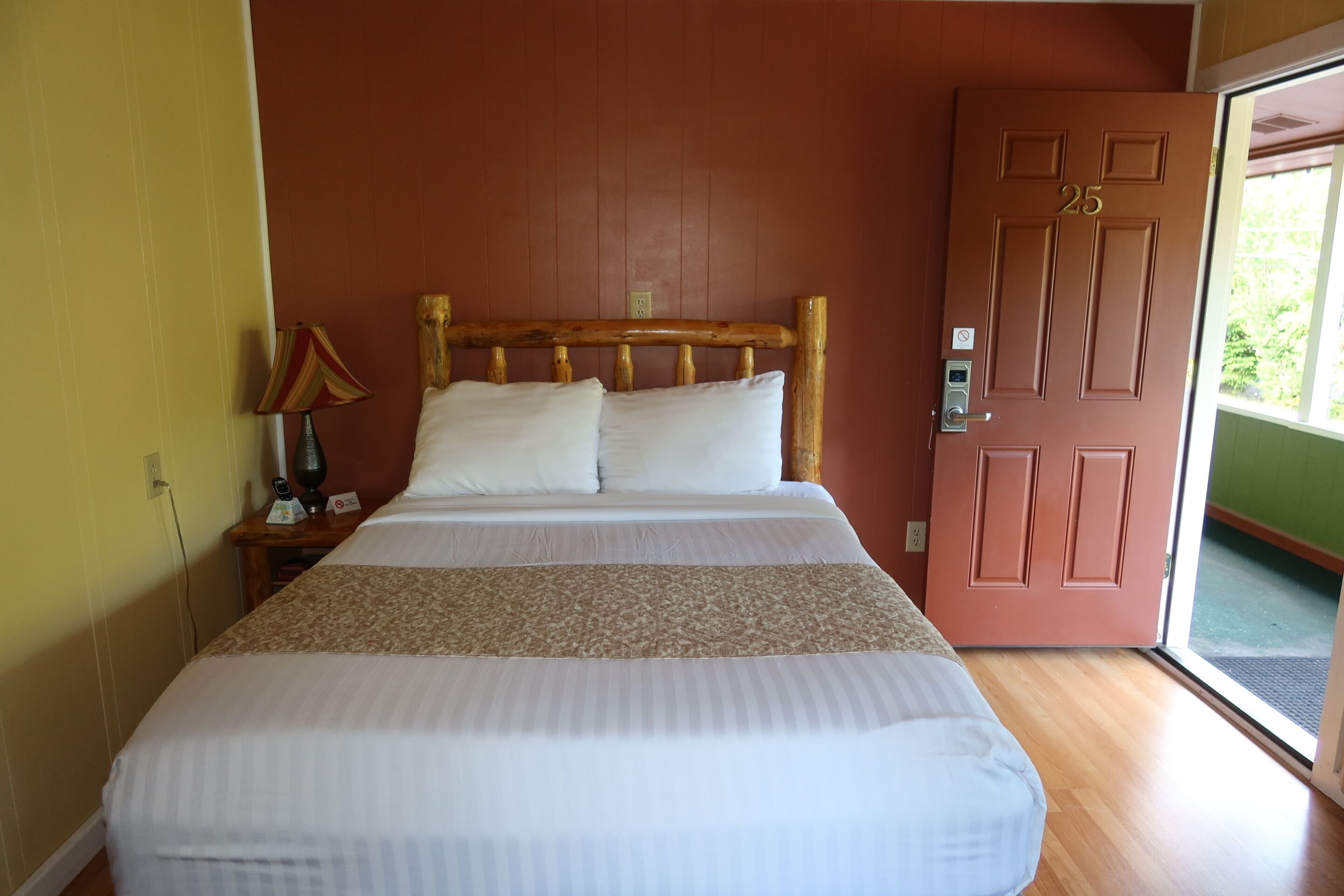 Een net opgemaakt bed met een houten hoofdbord, een bedlampje en een deur met een nummer.