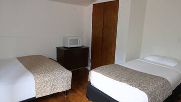 Quarto com duas camas, um guarda-roupa de madeira, uma mesinha com micro-ondas e uma porta.