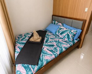 Un lit simple avec une couvre-lit ornée d’un motif de feuilles tropicales, un oreiller noir, un oreiller bleu et une serviette beige.