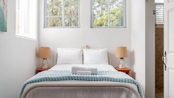 A bedroom with a bed, two bedside lamps, a window with blinds, and a door leading to another room.