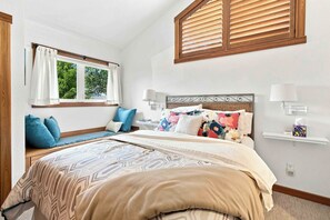 Een slaapkamer met een bed, een raam met jaloezieën, een nachtkastje met een lamp en een wandplank met een zakdoos.