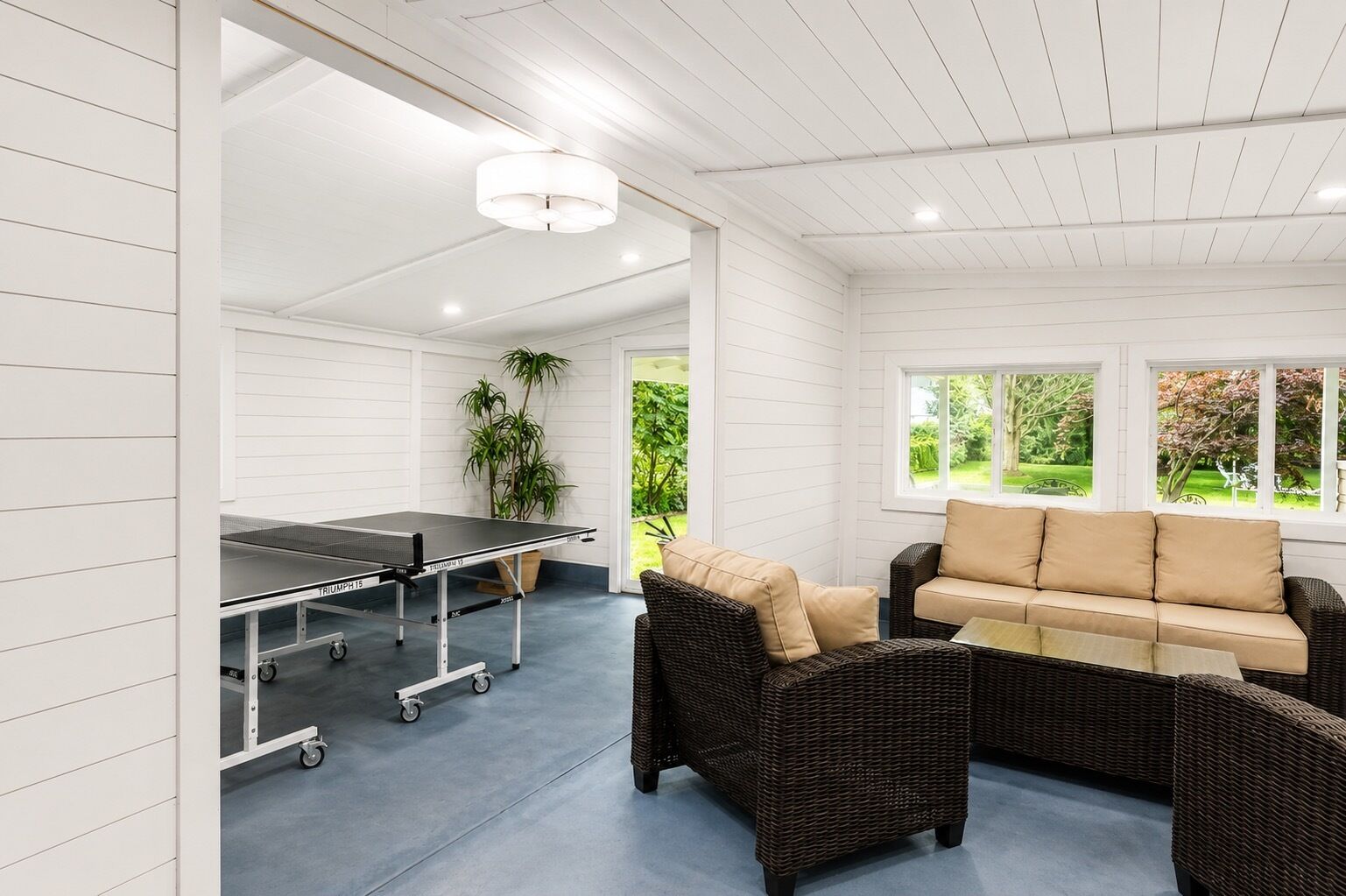 A room with a ping pong table, wicker furniture, and a view of greenery outside.