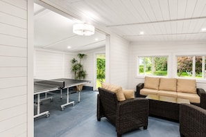 A room with a ping pong table, wicker furniture, and a view of greenery outside.
