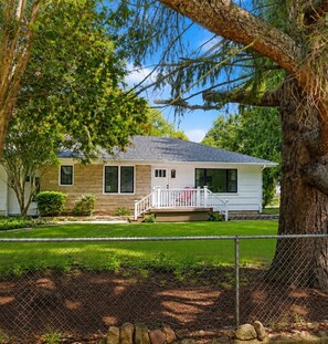 Ein eingeschossiges Haus mit Veranda, einem eingezäunten Garten und einem großen Baum im Vordergrund.