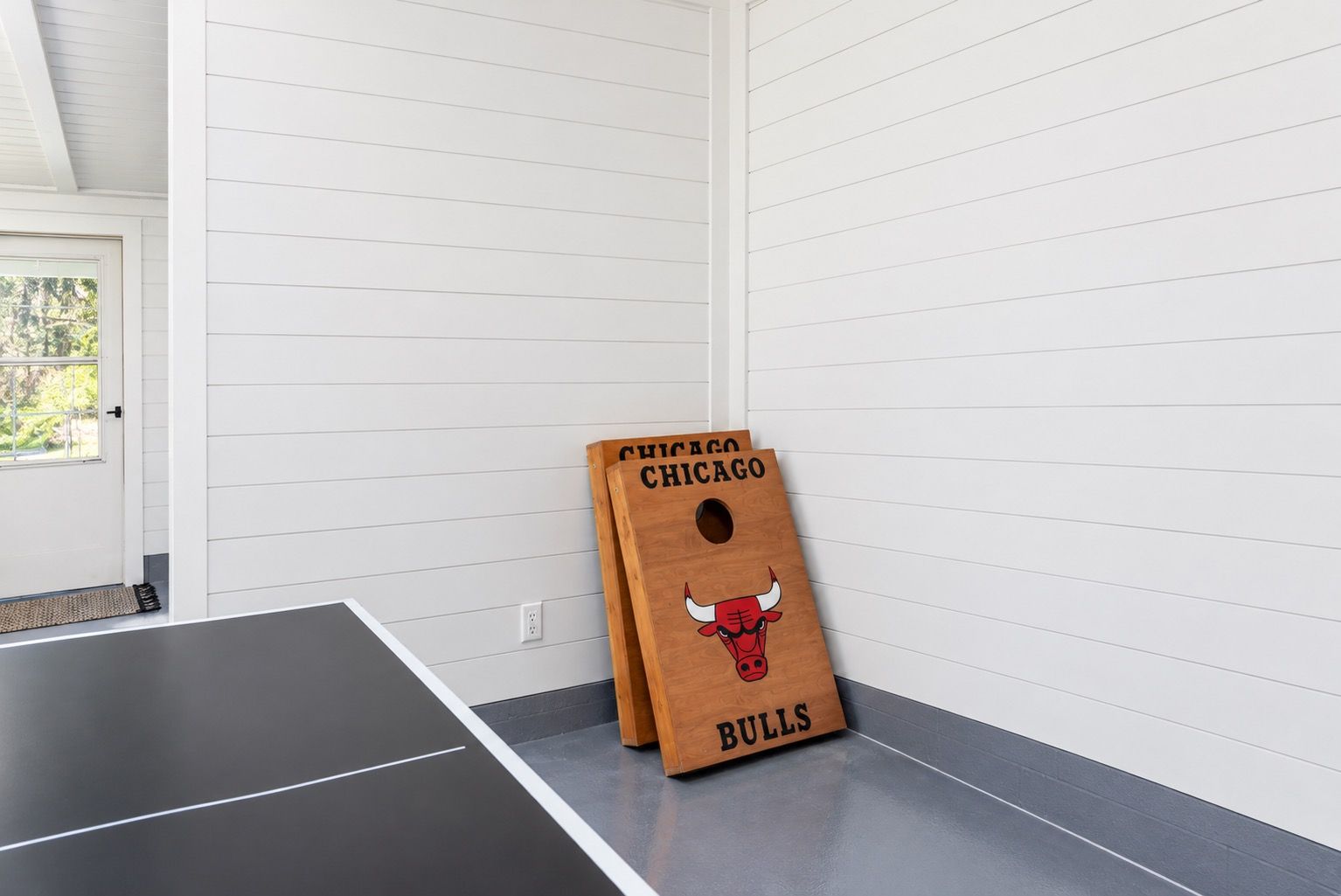 Une table de ping-pong dans une pièce aux murs recouverts de panneaux blancs, avec une porte donnant sur un paysage verdoyant.