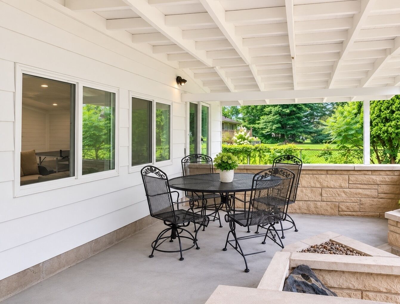 A covered patio with a round table and chairs, large windows, and a stone fire feature.