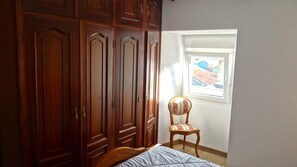 Une chambre à coucher avec un lit, une armoire en bois, une chaise et une fenêtre donnant sur des immeubles.