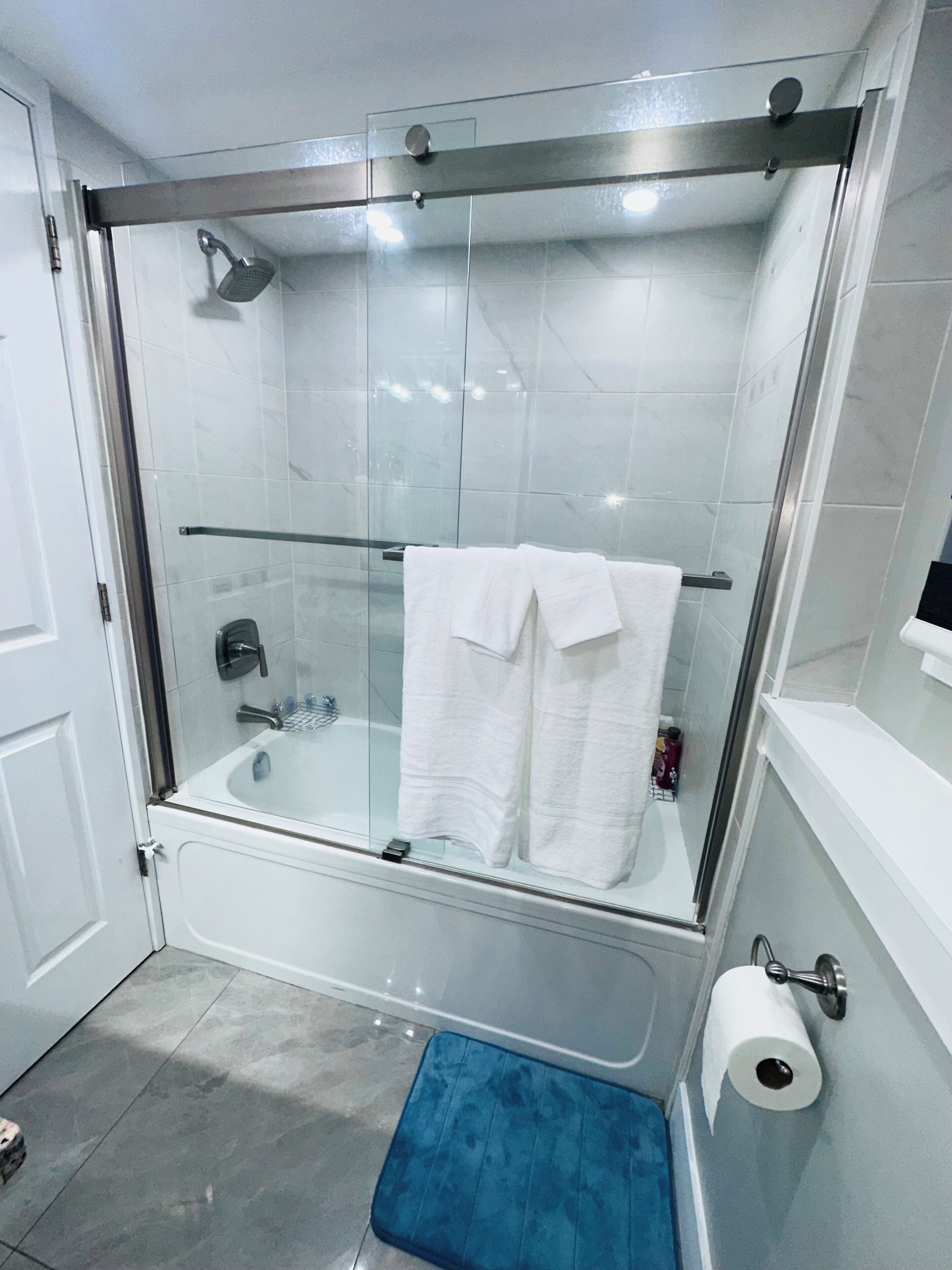 A modern bathroom with a glass-enclosed shower, a bathtub, and a blue bath mat.