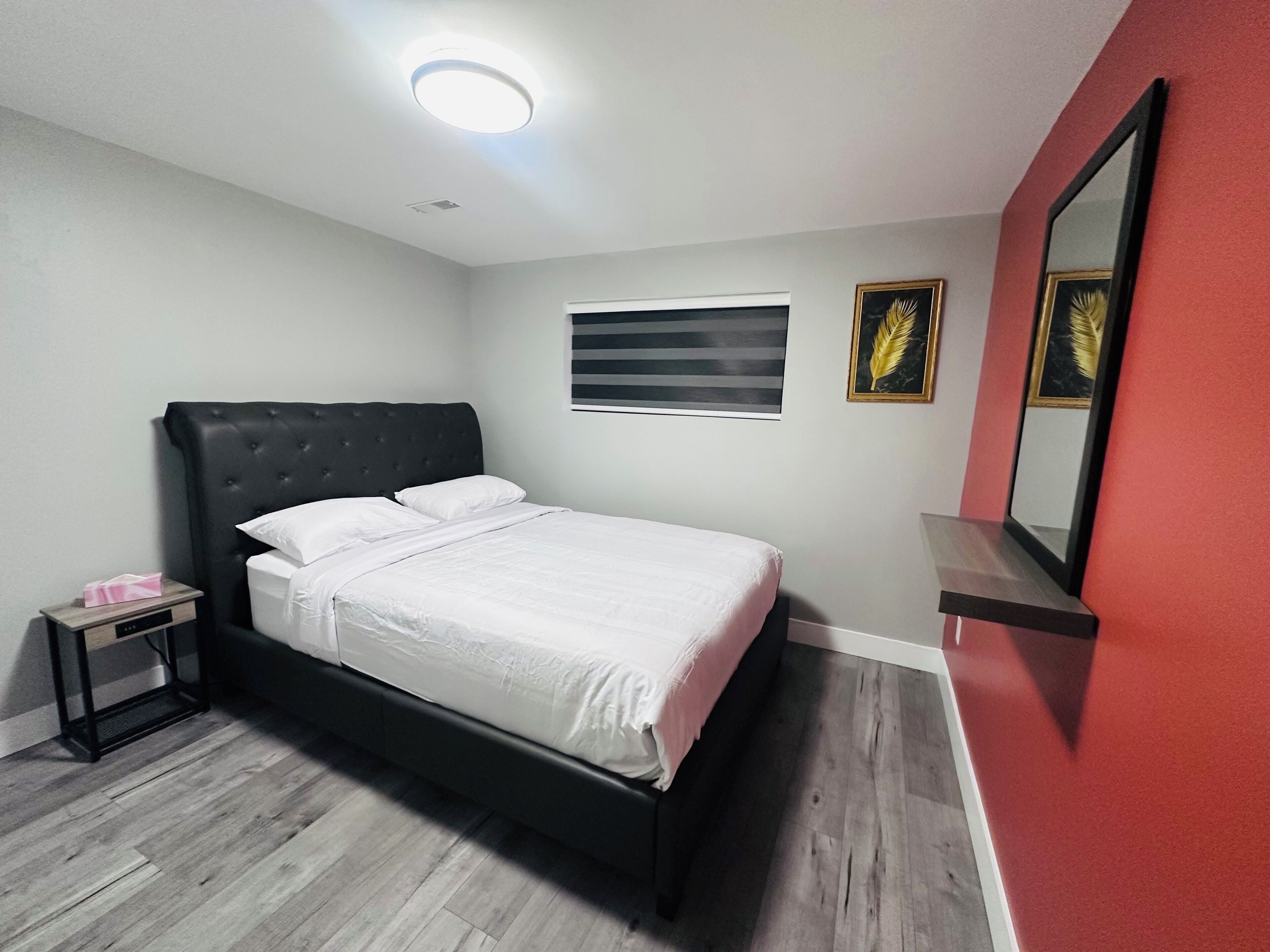 Een slaapkamer met een bed, nachtkastje, spiegel en raam met jaloezieën.
