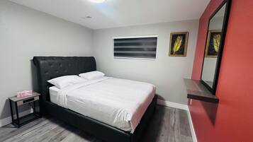 Een slaapkamer met een bed, nachtkastje, spiegel en raam met jaloezieën.