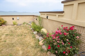 A patio area with a brick wall, a grassy lawn, and a flower bed with pink flowers. - New! Stunning Lake & Mountain Views, Sauna, Backyard, Walk to Downtown Polson! (Polson)